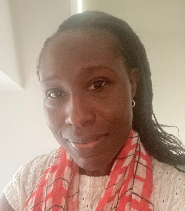 Portrait of a dark brown woman with braided hair, small earrings, wearing a white top and red scarf.